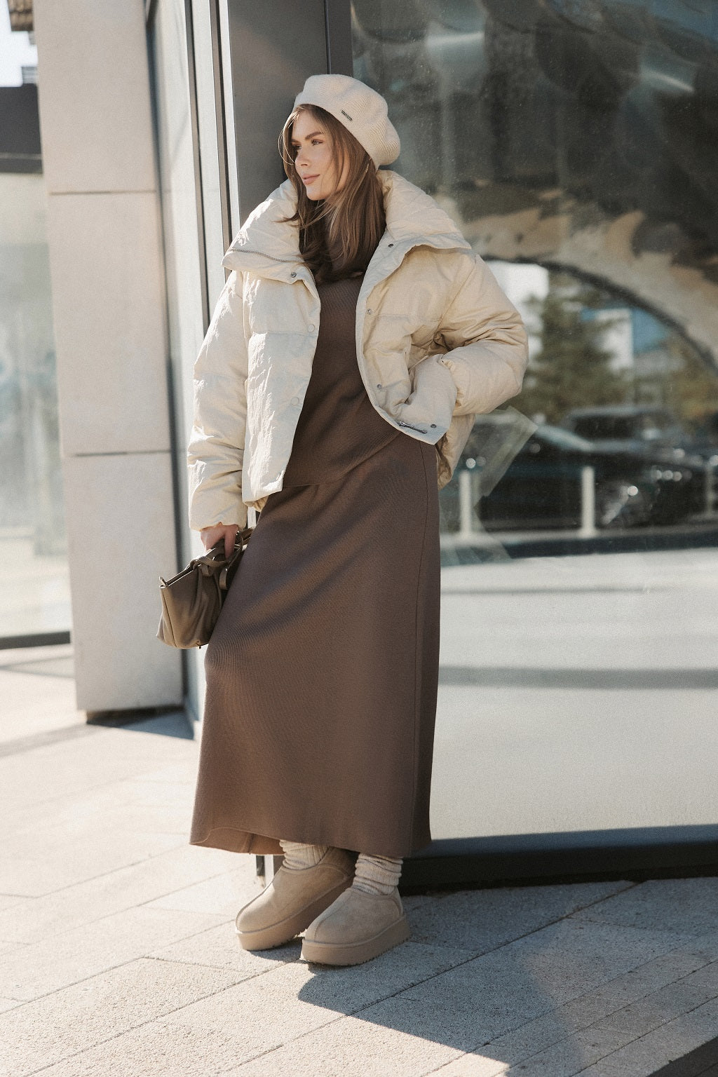 Woman in a winter outfit with a puffer jacket and brown dress standing outside a building.