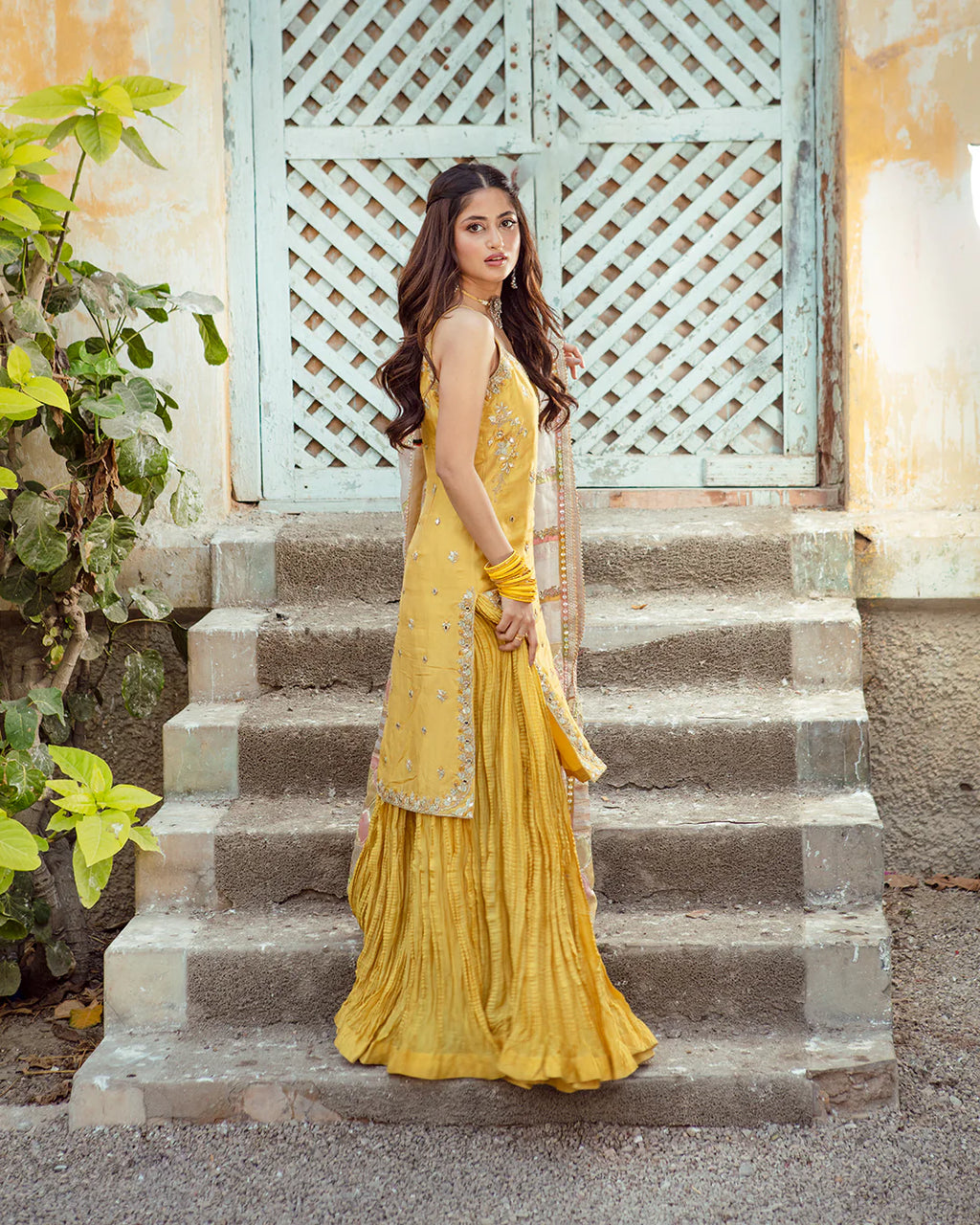 Woman in a yellow traditional outfit standing on stone steps with a decorative white lattice in the background.