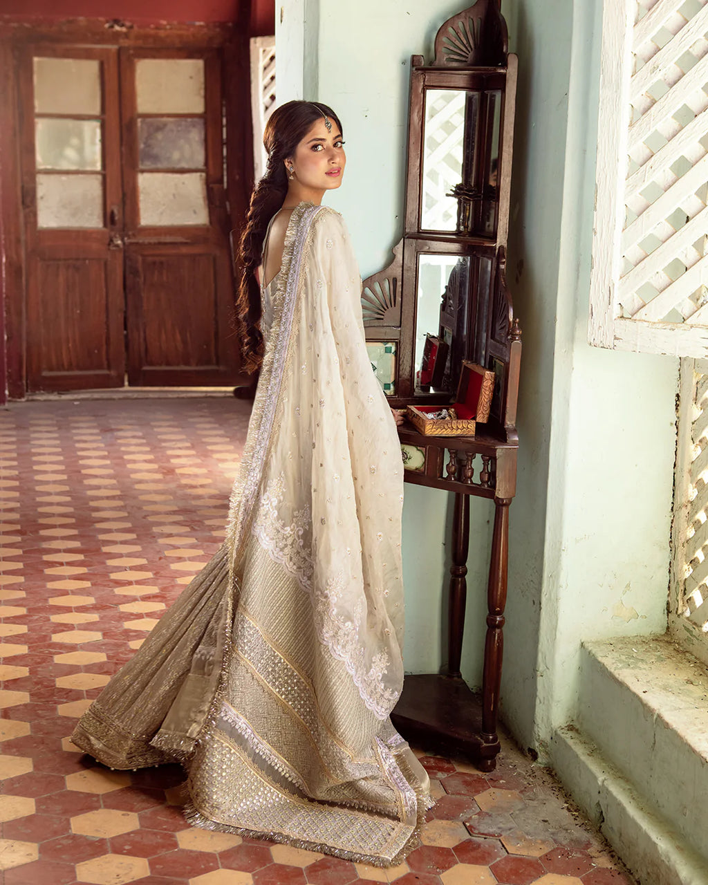 Woman in a traditional outfit standing in a room with wooden furniture and a patterned floor.