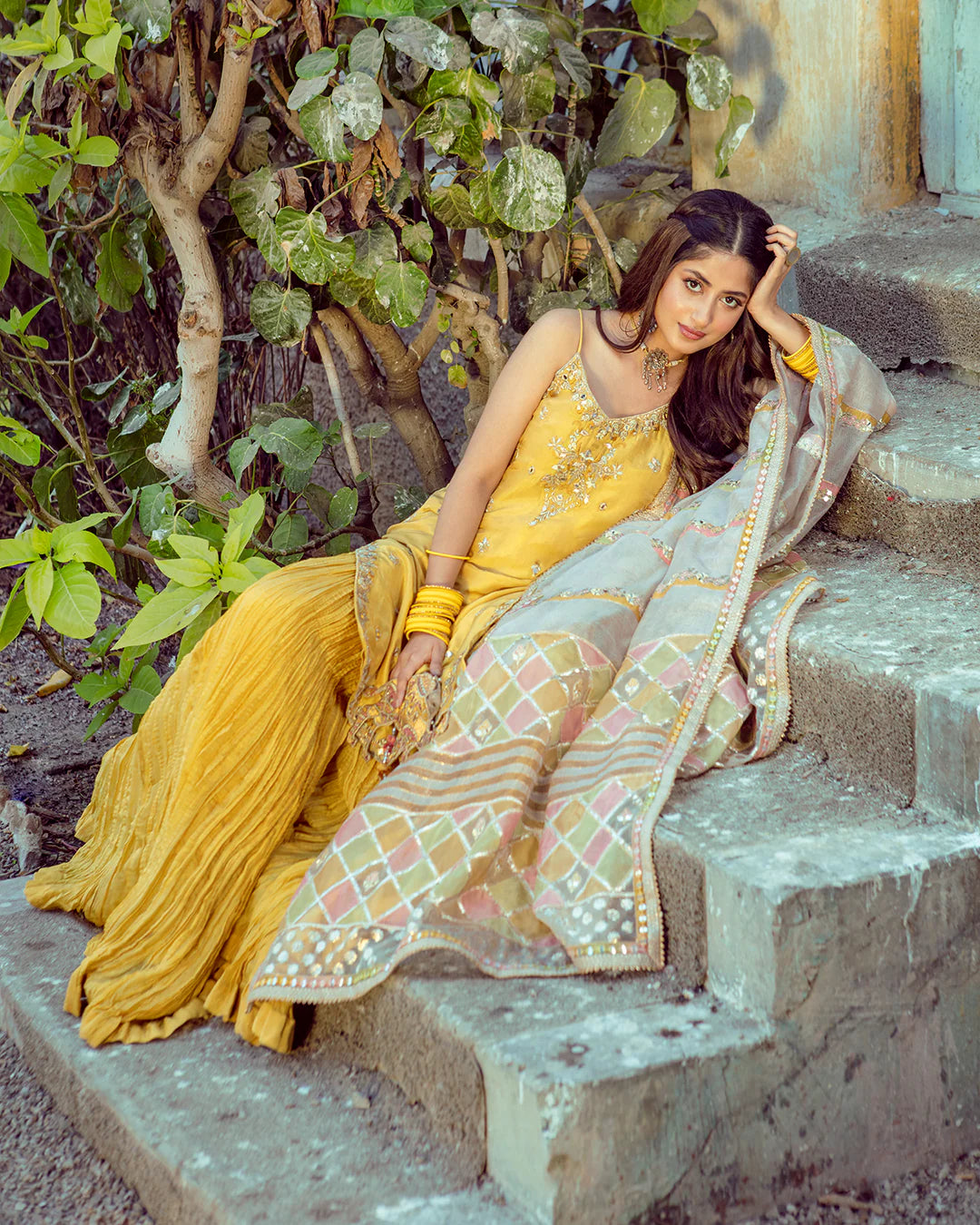 Woman in traditional yellow and patterned saree sitting on steps with greenery in the background