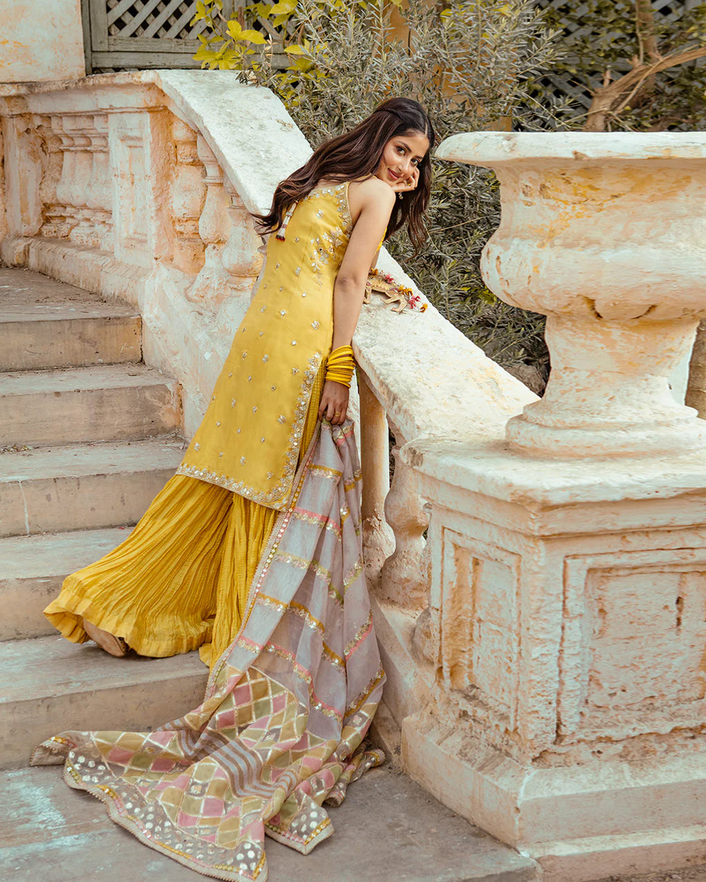 Woman in a yellow traditional outfit standing on stone steps with a decorative stone structure in the background.
