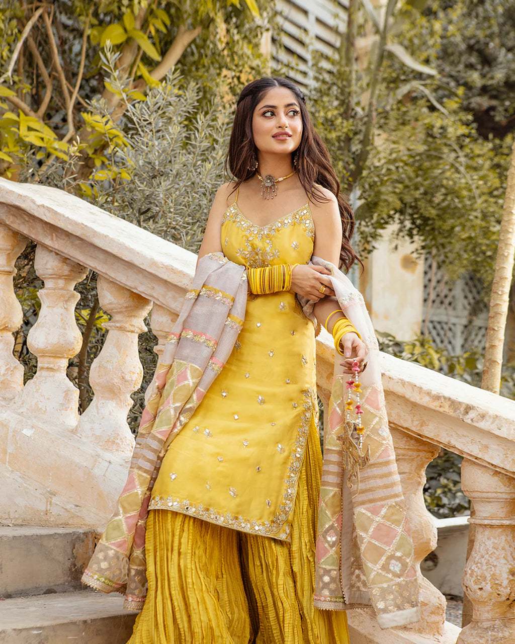 Woman in a yellow traditional outfit standing outdoors with greenery in the background