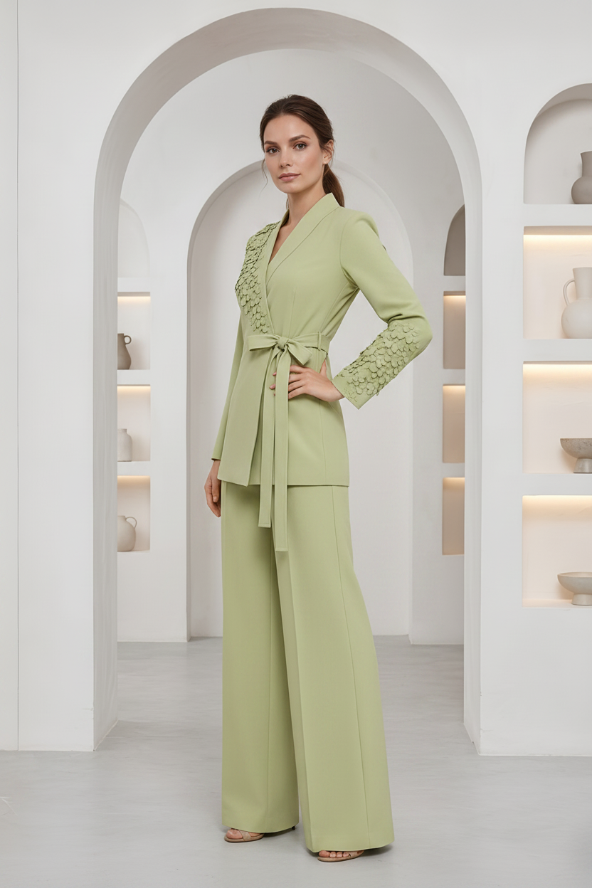 A woman models a sage green two-piece set consisting of a wrap-style long-sleeve top with lace detailing on the front and sleeves, paired with matching wide-leg trousers, in a bright white arched interior setting.