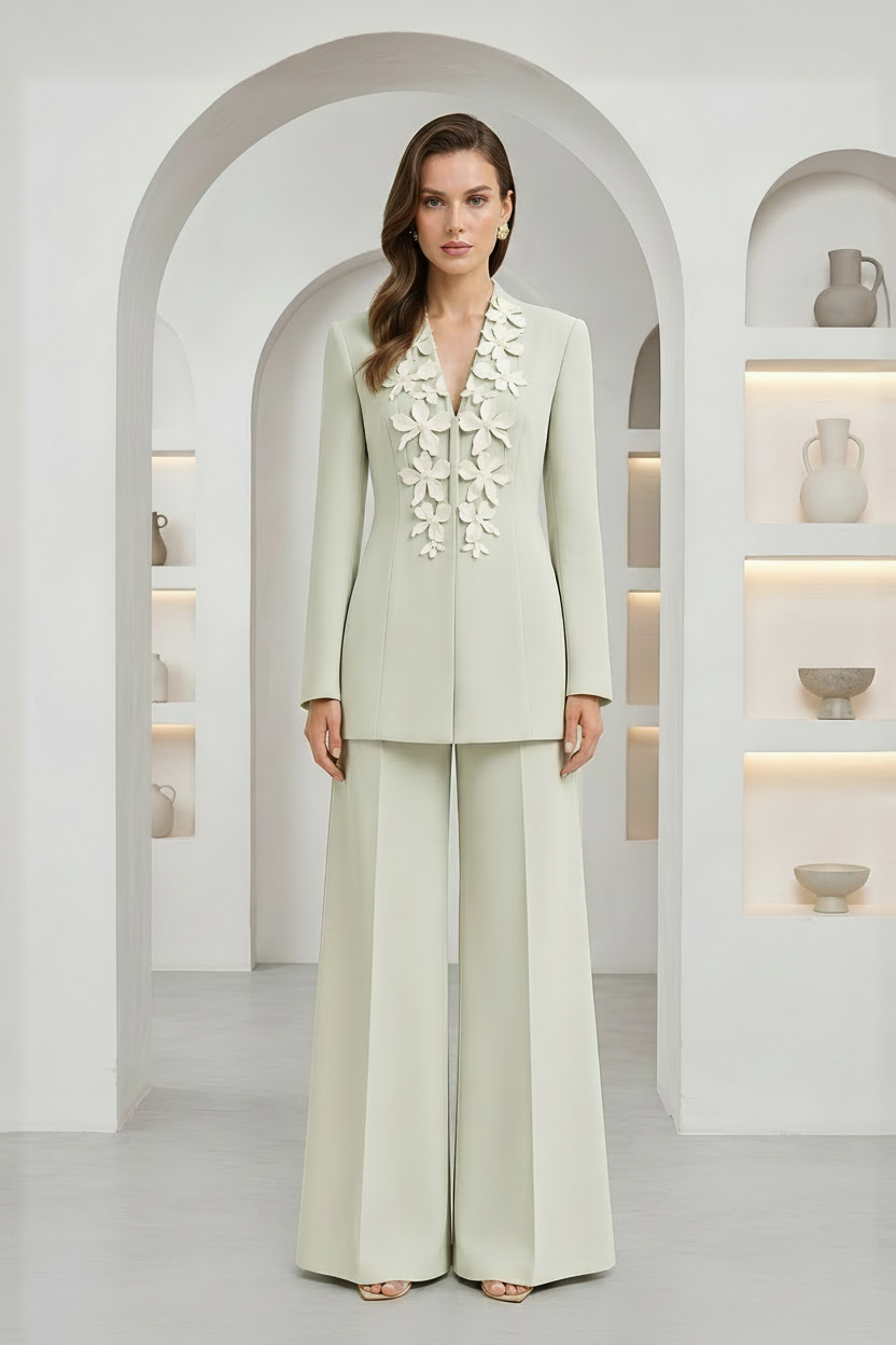 A woman models a light sage green two-piece suit with wide-leg trousers and a matching blazer featuring intricate floral embroidery on the chest, set against a minimalist white arched interior.