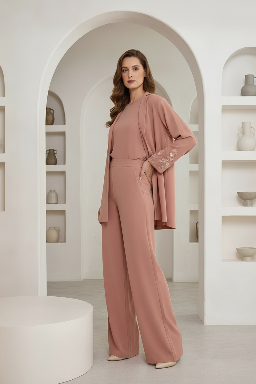 A woman models a dusty rose two-piece outfit consisting of a wide-leg jumpsuit and a matching draped jacket with embroidered cuffs, set against a minimalist white arched interior with shelves.