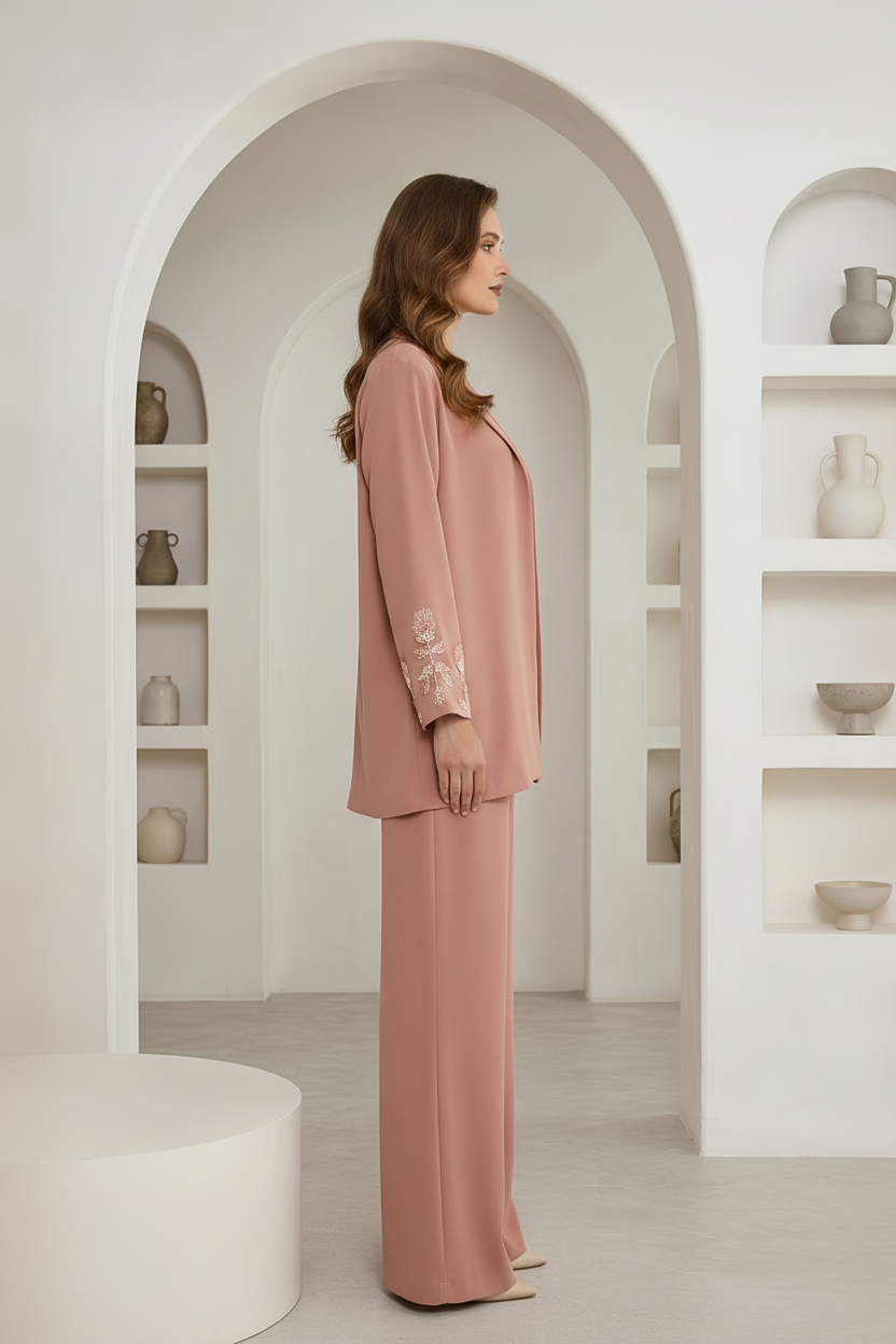 A woman stands in profile wearing a dusty pink two-piece suit with a long-sleeve blazer and wide-leg trousers. The blazer has white floral embroidery on the sleeve.