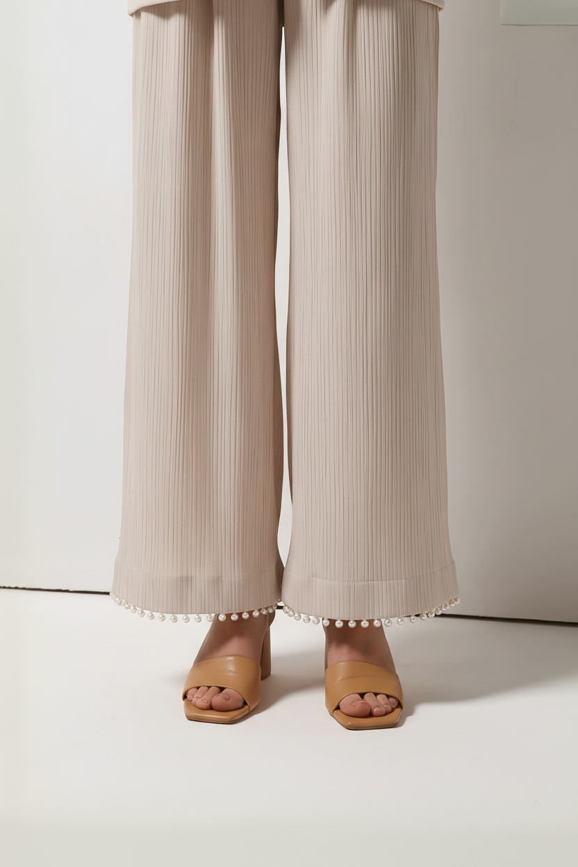 A person wearing wide-leg pleated pants in a light beige/taupe color with a row of small white pearls along the hem, paired with tan heeled sandals, shown from the waist down against a plain light background.