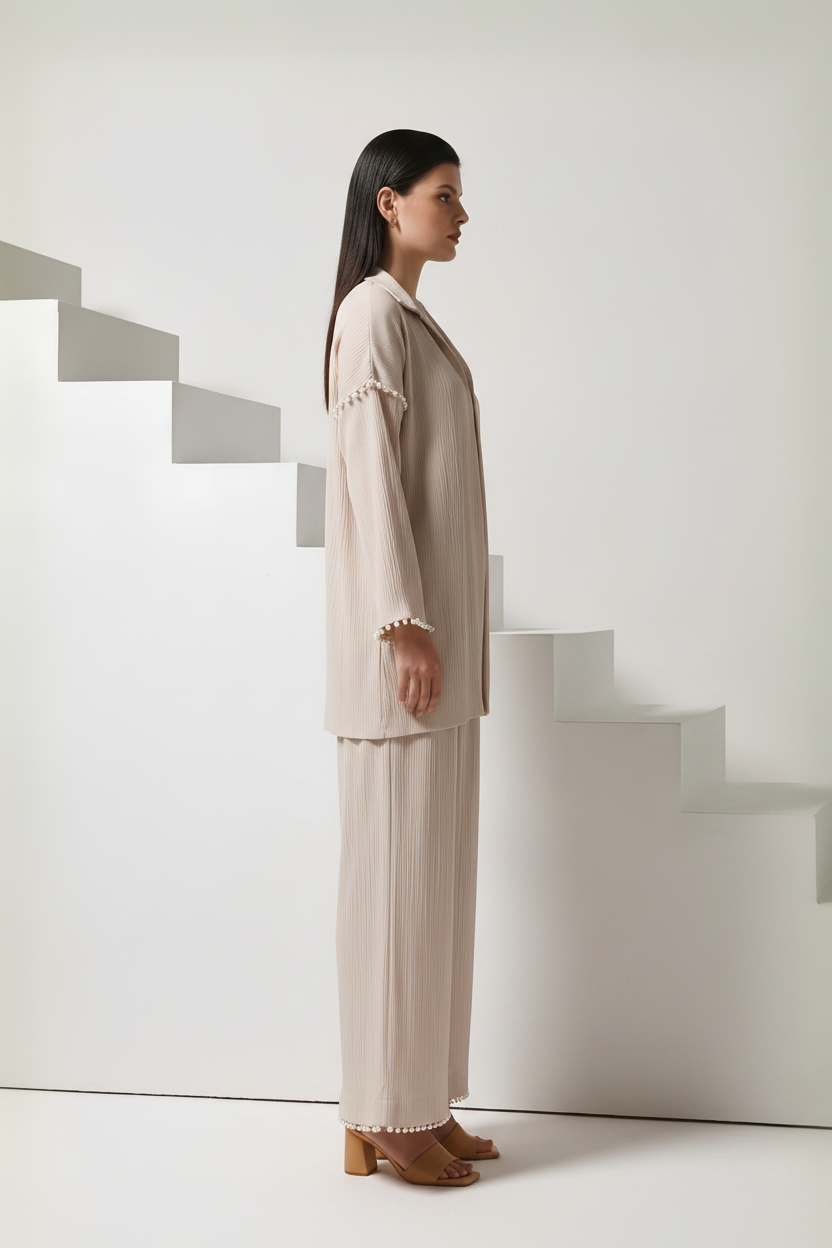 A woman stands in profile wearing a light beige two-piece set with a cropped blazer and wide-leg pleated pants, styled with brown heeled sandals. She is posed in front of minimalist white geometric stairs.