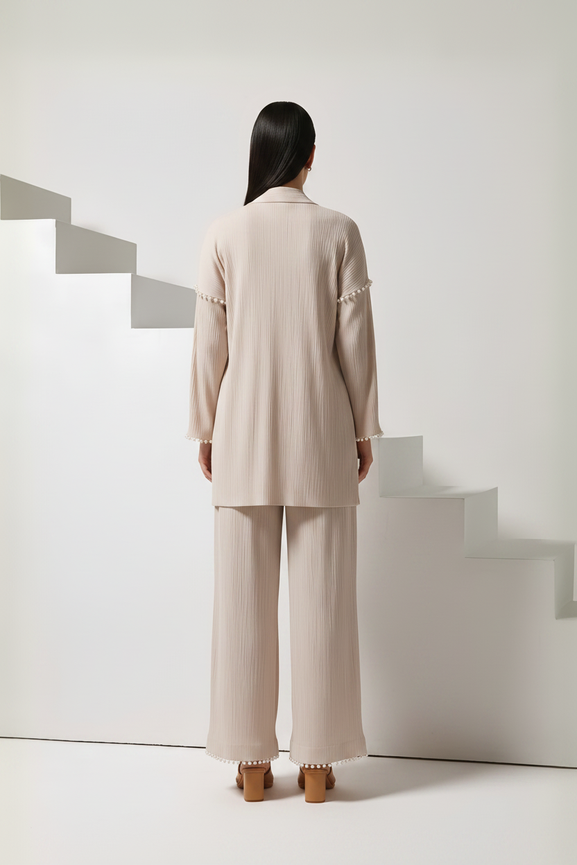 A model viewed from behind wearing a light beige ribbed two-piece set consisting of a long-sleeve blazer with decorative ruffle trim at the cuffs and matching wide-leg trousers, set against a minimalist white background with geometric steps.