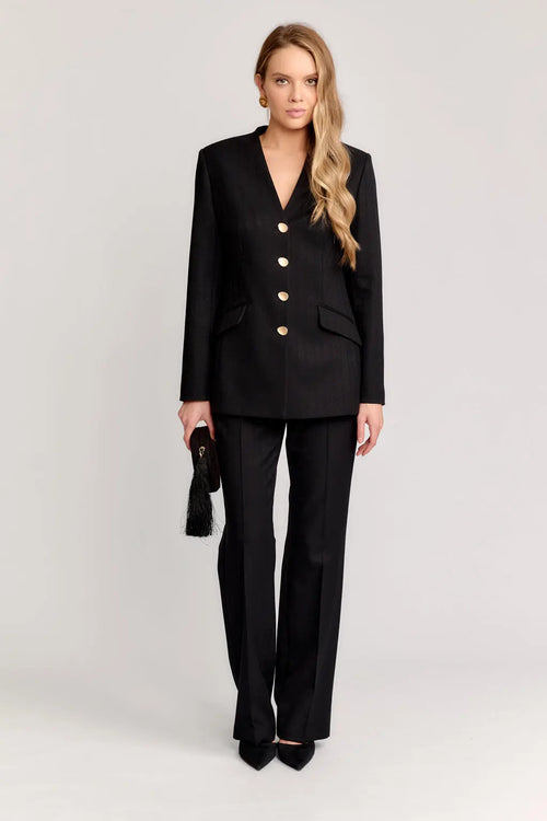 Woman wearing a black suit with a white background Front view of the DEITY Obsidian sackcloth jacket showing the lapel-less V-neck.