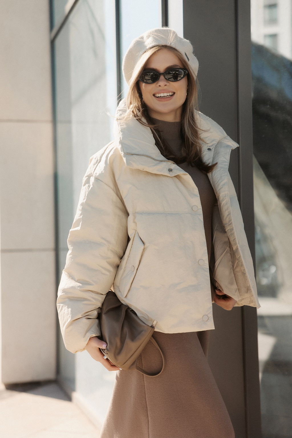 Woman wearing a beige puffer jacket and sunglasses, standing against a building.