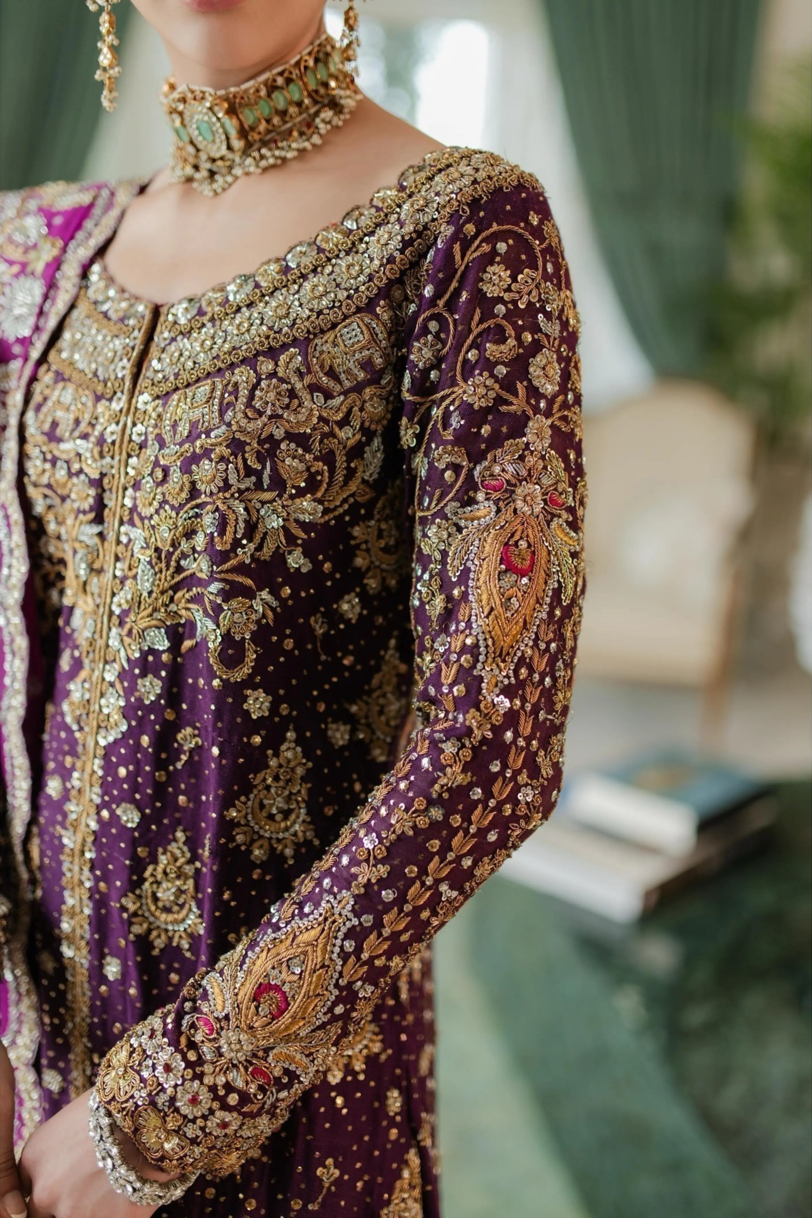 A close-up of a person wearing an ornate deep purple embroidered traditional outfit with extensive gold detailing, a matching necklace, and earrings in a softly blurred indoor setting.