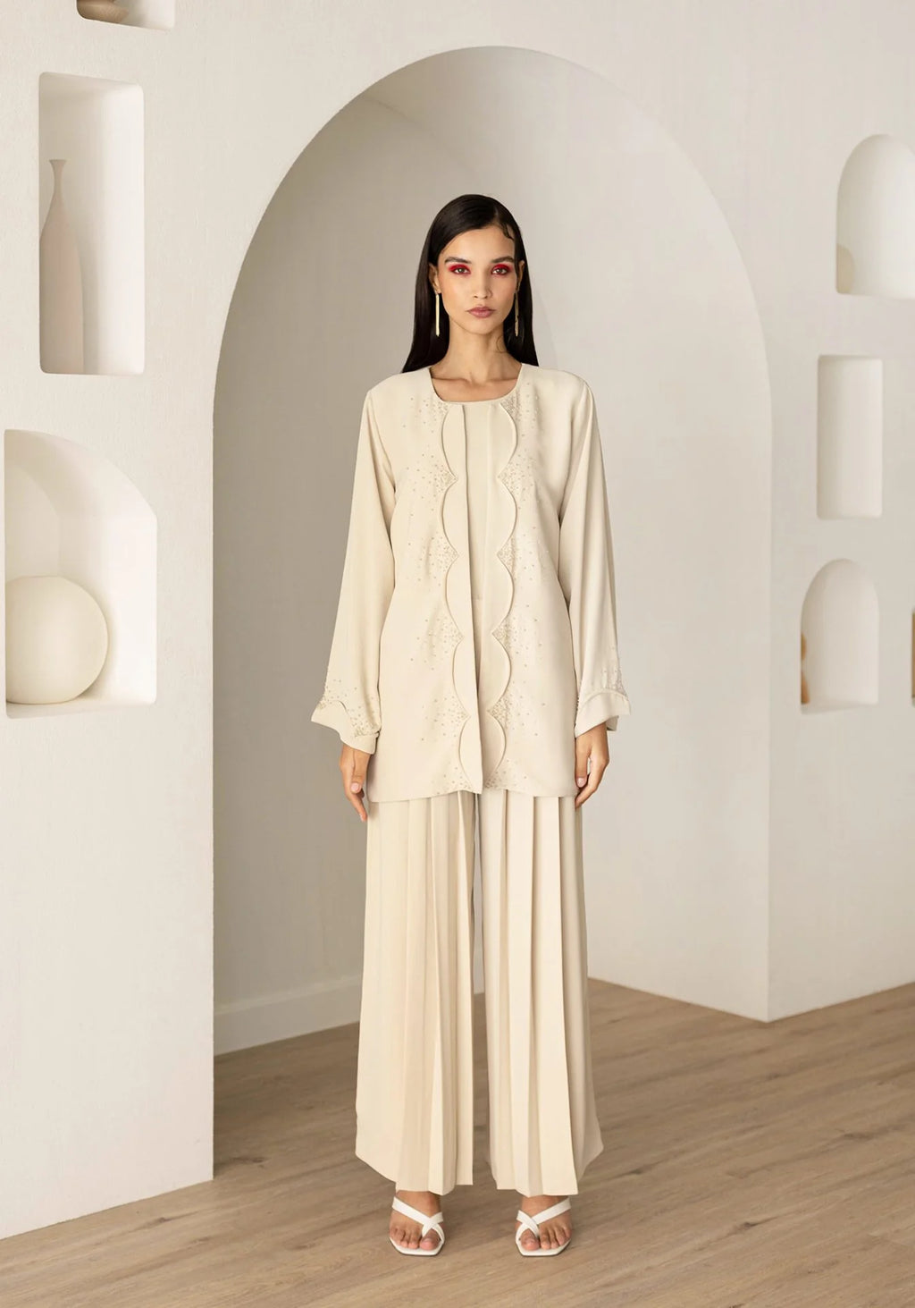 A woman stands in a minimalist cream-colored room with arched alcoves and light wood flooring, wearing a matching cream-colored two-piece outfit consisting of a long-sleeve embroidered tunic and wide-leg trousers.