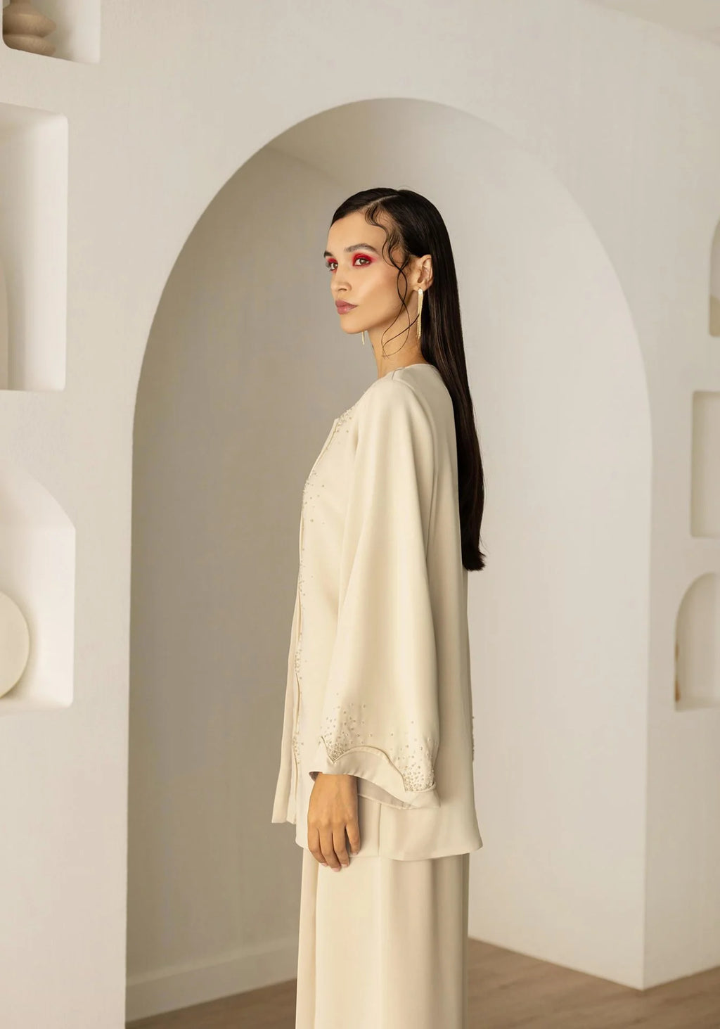 A woman stands in profile wearing a cream-colored traditional outfit with wide sleeves and embroidered details, set against a minimalist white arched wall.