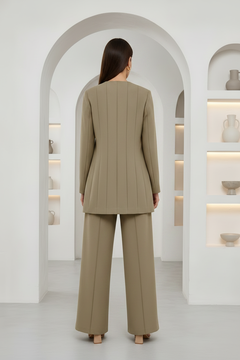 A model viewed from behind wearing a matching beige pleated blazer and wide-leg trousers set, standing in a bright, modern white room with arched alcoves and built-in shelves.