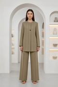 A model stands in a minimalist white architectural setting with arched passageways and built-in shelves, wearing a matching taupe-beige two-piece suit consisting of a long-sleeve pleated jacket with a single button and wide-leg trousers.