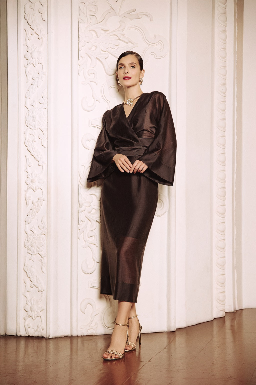 Woman in a dark brown dress standing against a textured white wall.