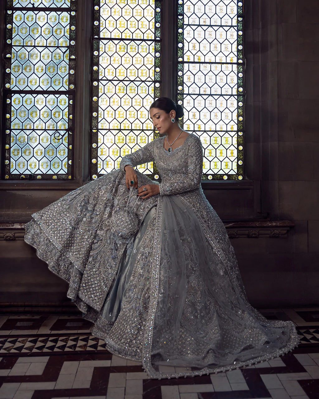 Woman in a silver embroidered traditional outfit sitting in front of decorative windows.