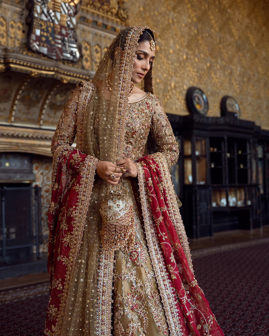 Woman in a traditional embroidered outfit with a red and gold color scheme, standing in an ornate room.