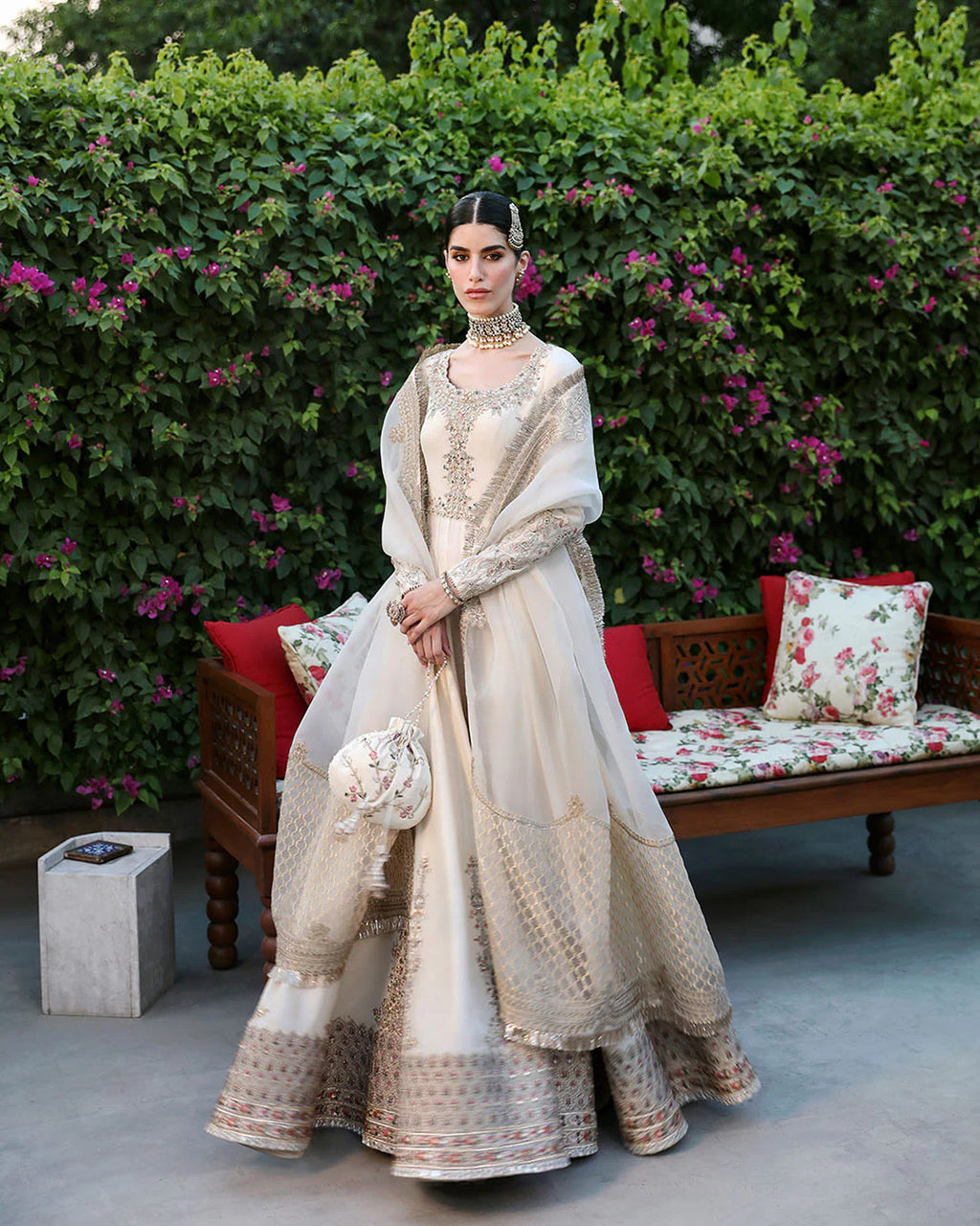 Woman in a traditional outfit standing in front of a floral-patterned bench with greenery in the background