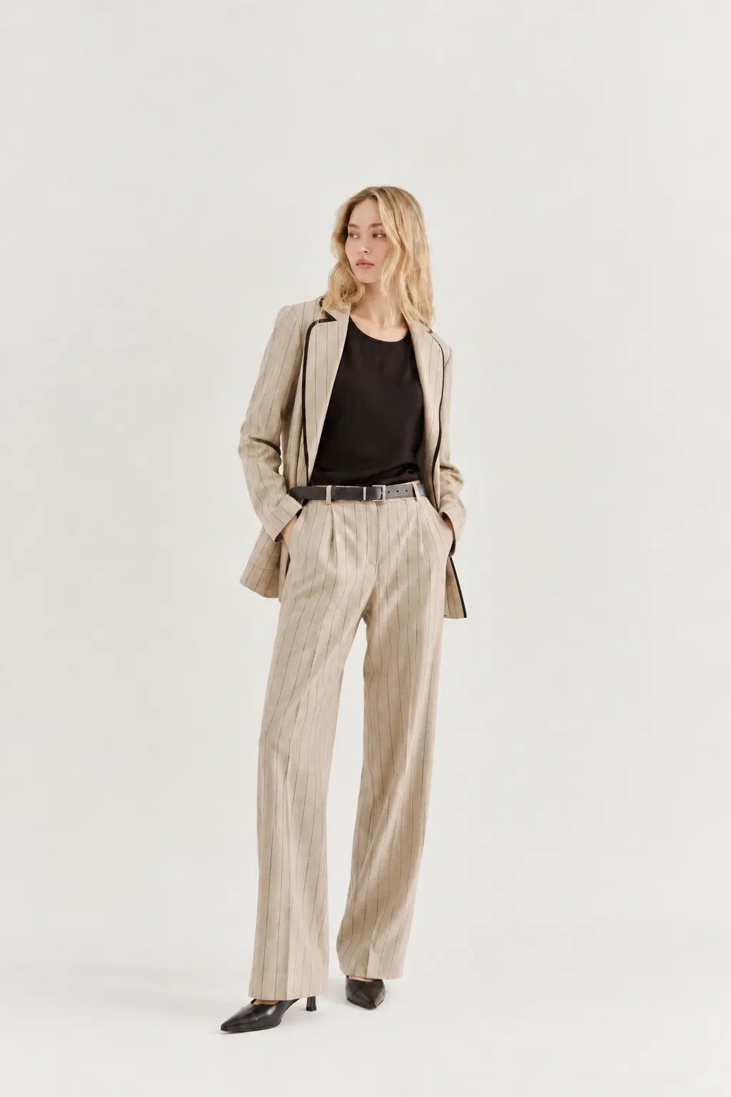 A woman modeling a beige pinstripe blazer and matching wide-leg trousers with a black top and black shoes, standing against a plain white studio background.