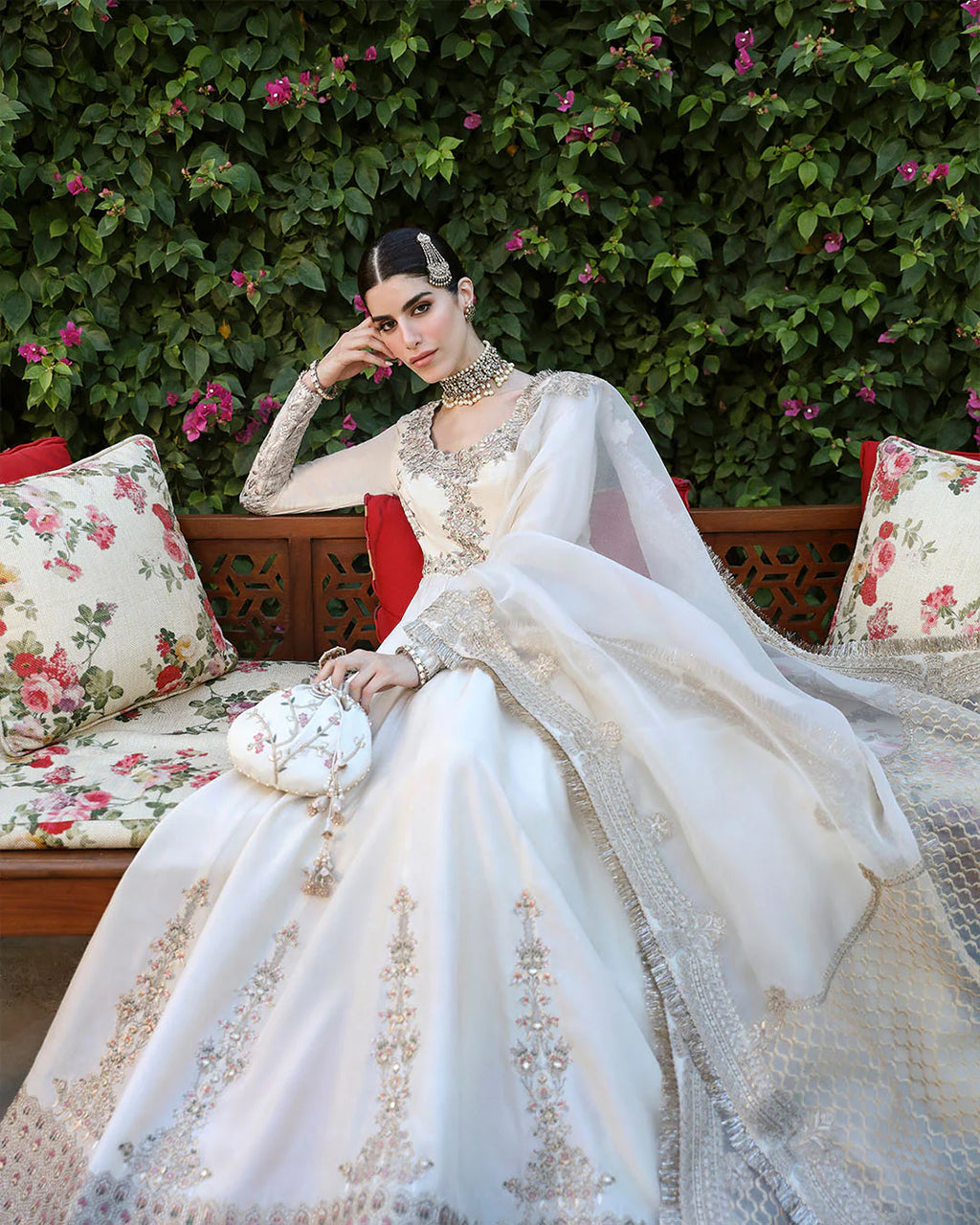 Woman in an ornate white dress sitting on a floral-patterned couch with green foliage in the background.
