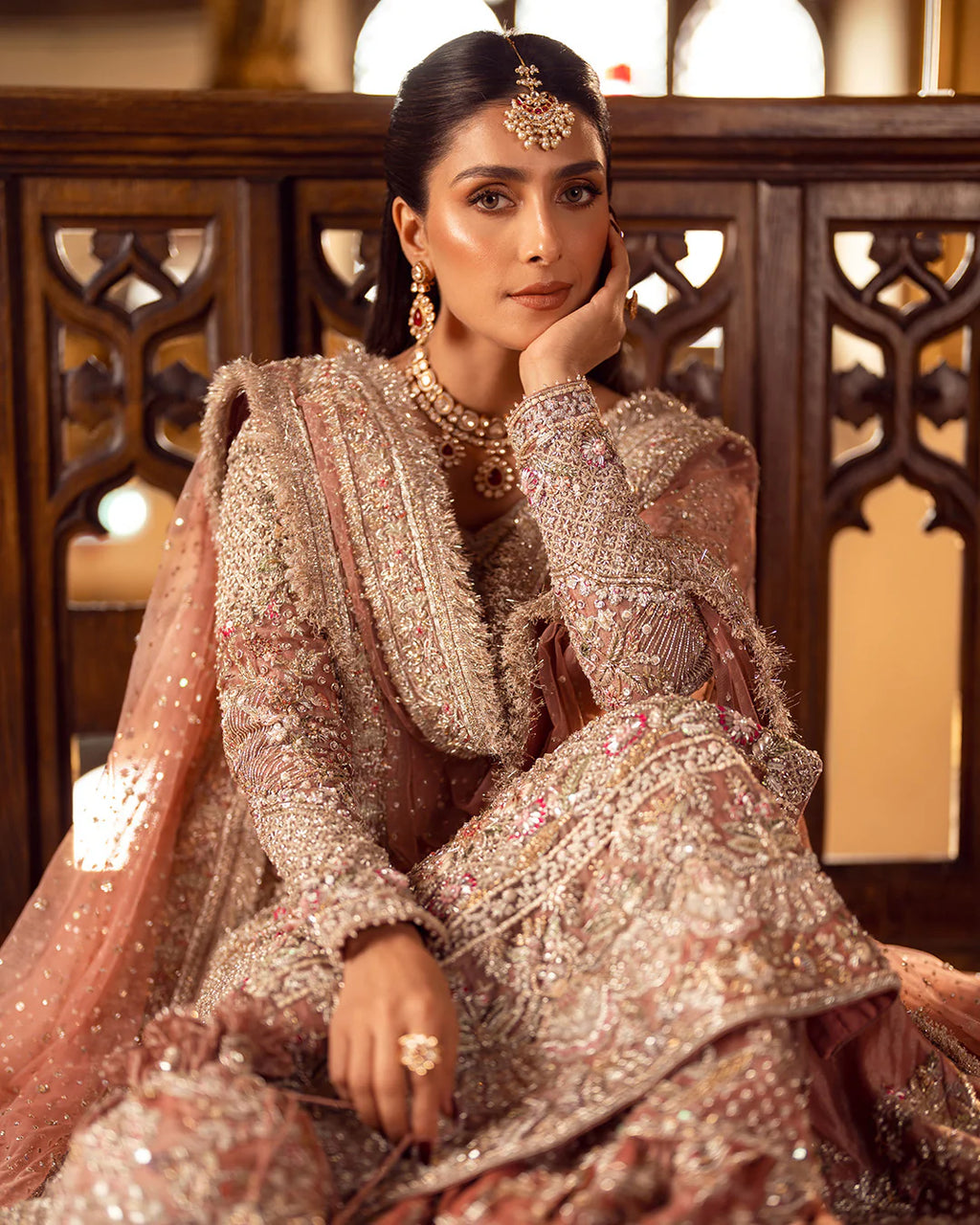 Woman in traditional embroidered outfit with jewelry, sitting against a decorative wooden panel.