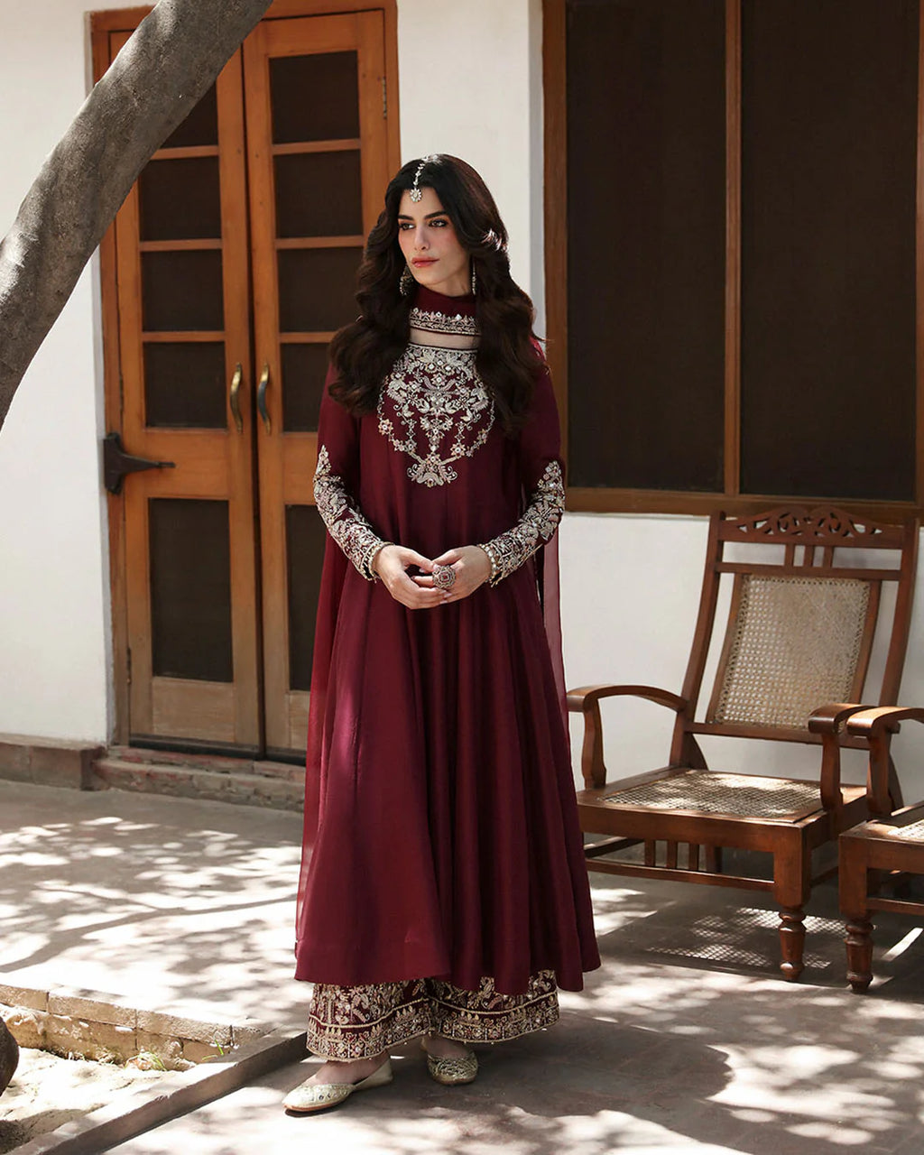 Woman in a maroon traditional outfit with intricate patterns standing in a sunlit room.