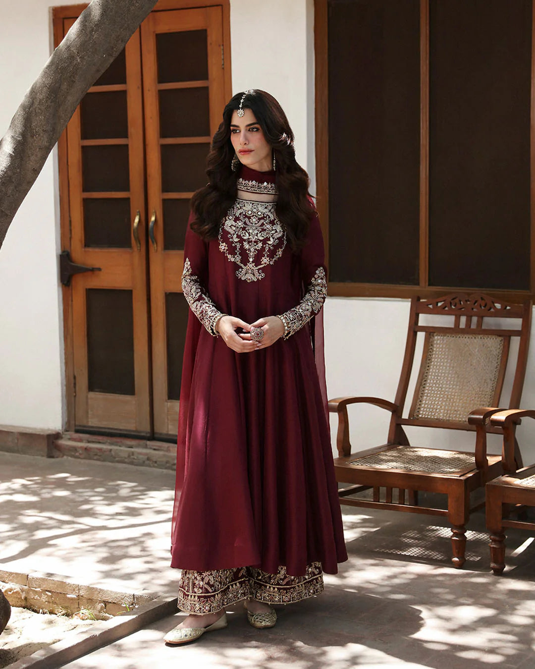 Woman in a maroon traditional outfit with intricate patterns standing in a sunlit room.