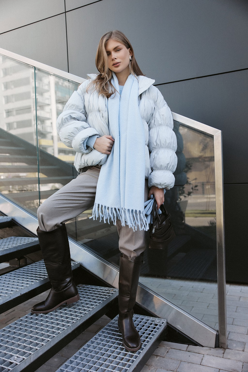 Woman in a stylish outfit with a puffer jacket, scarf, and knee-high boots standing on metal stairs.