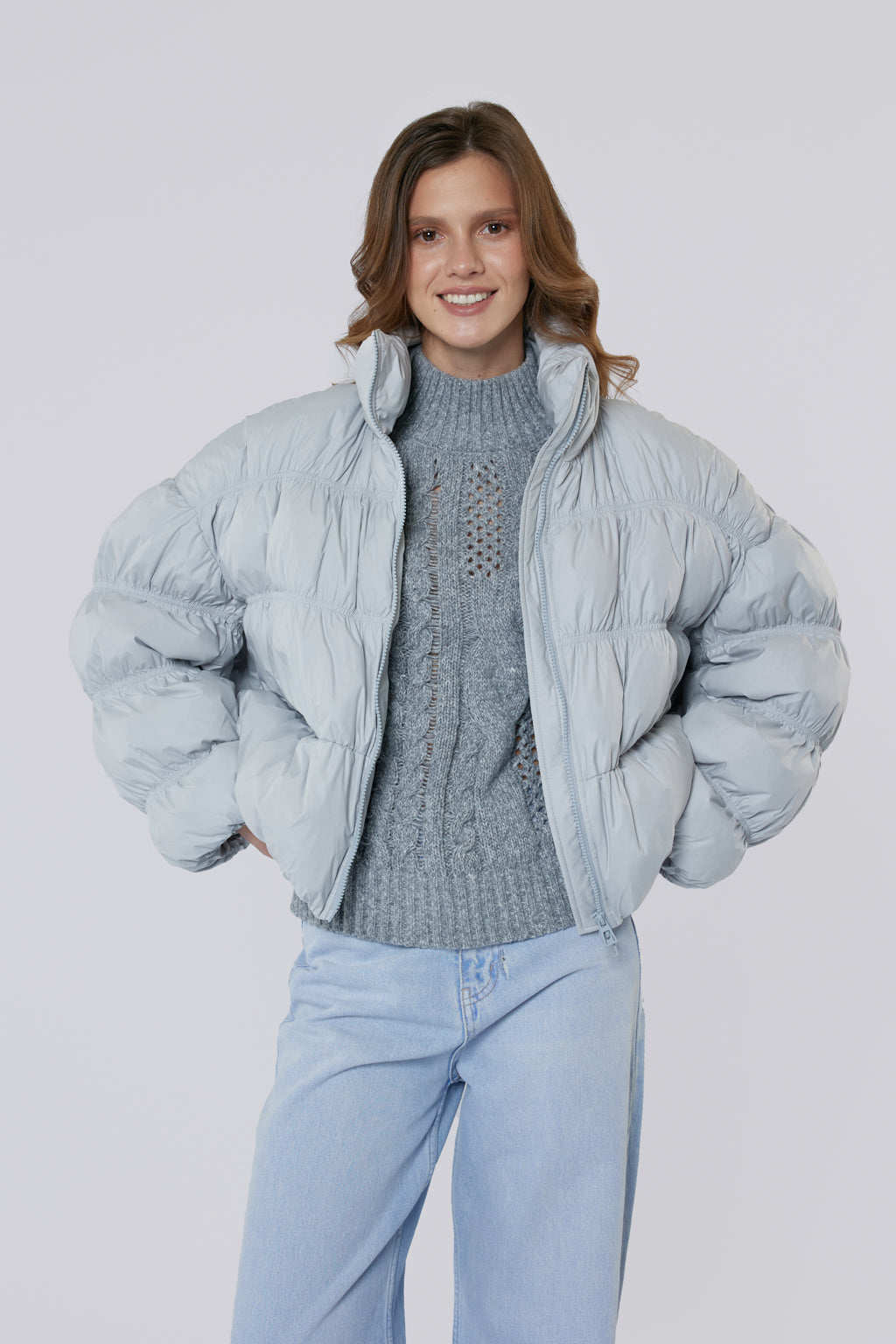 Woman wearing a light blue puffer jacket over a gray sweater and light blue jeans on a white background