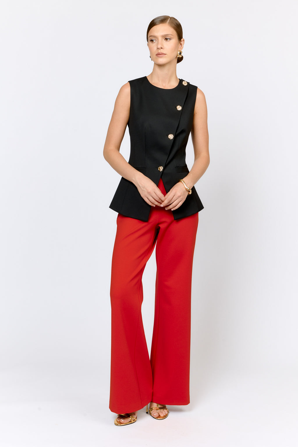 Woman wearing a black sleeveless top with gold buttons and red pants on a white background