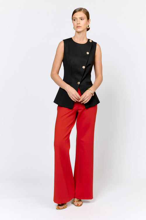 Woman wearing a black sleeveless top with gold buttons and red pants on a white background
