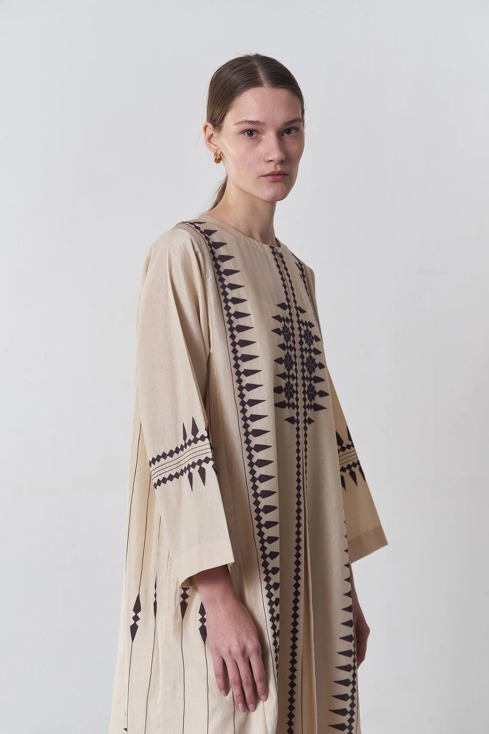 A floor-length beige Chanderi dress featuring vertical panels of ivory geometric tribal hand-cut appliqué on the bodice and hem.