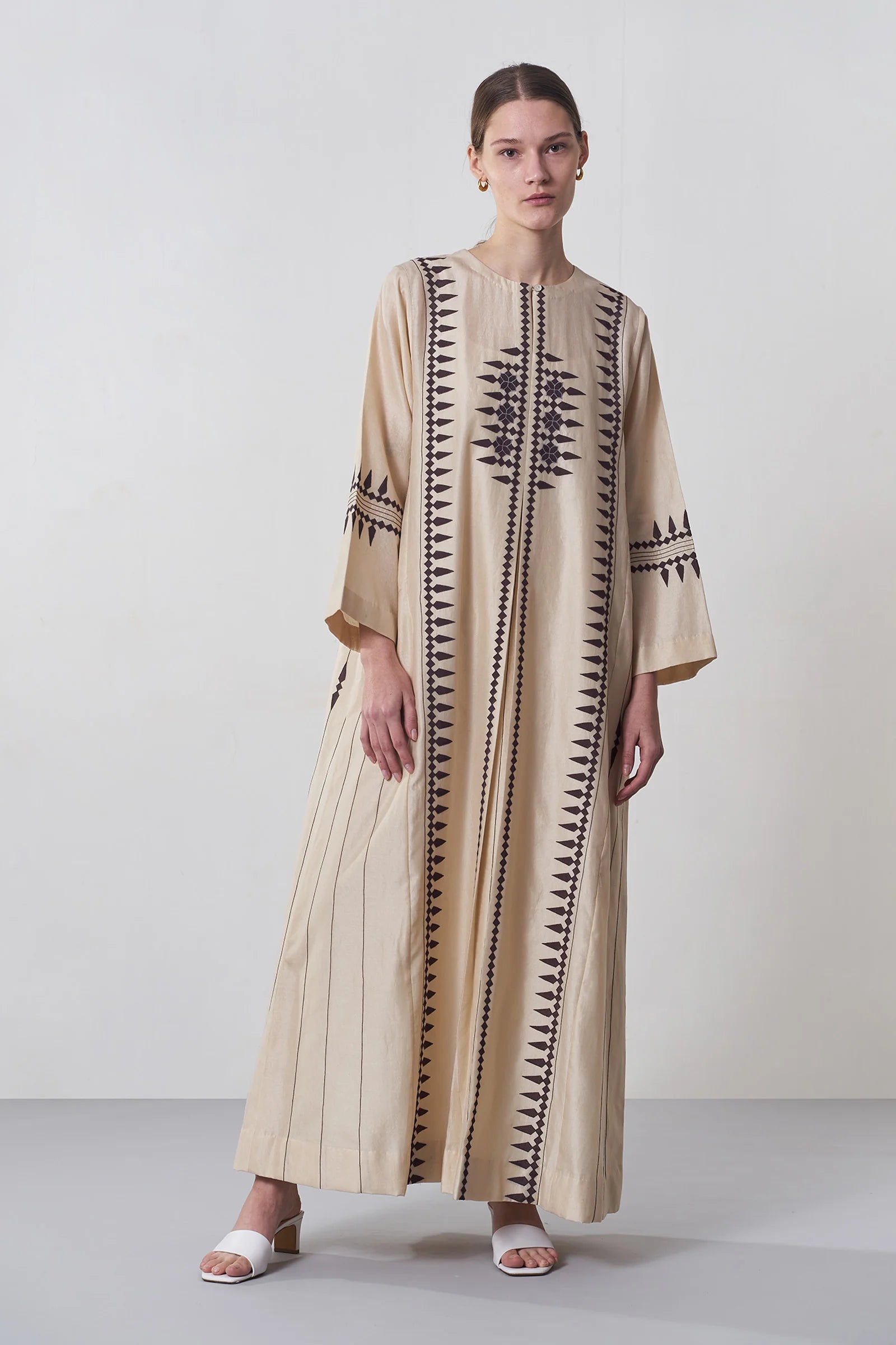 A floor-length beige Chanderi dress featuring vertical panels of ivory geometric tribal hand-cut appliqué on the bodice and hem.