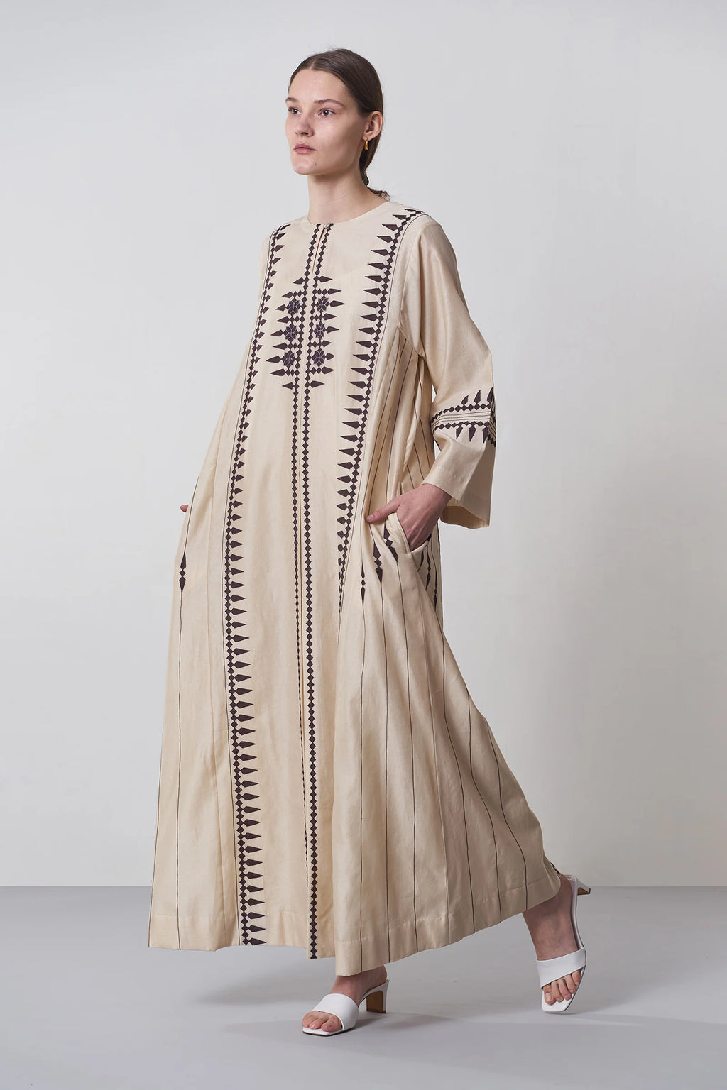 A floor-length beige Chanderi dress featuring vertical panels of ivory geometric tribal hand-cut appliqué on the bodice and hem.