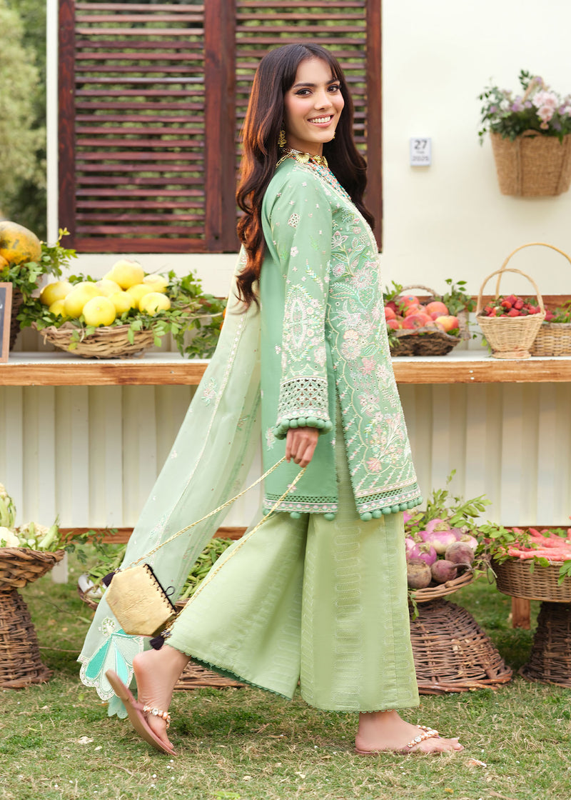 Woman in a light green traditional outfit standing outdoors with a table of fruits and flowers in the background.
