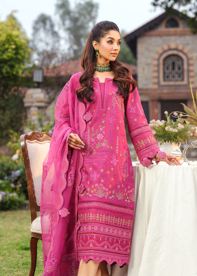 Woman in a pink embroidered outfit standing outdoors with a building and garden in the background