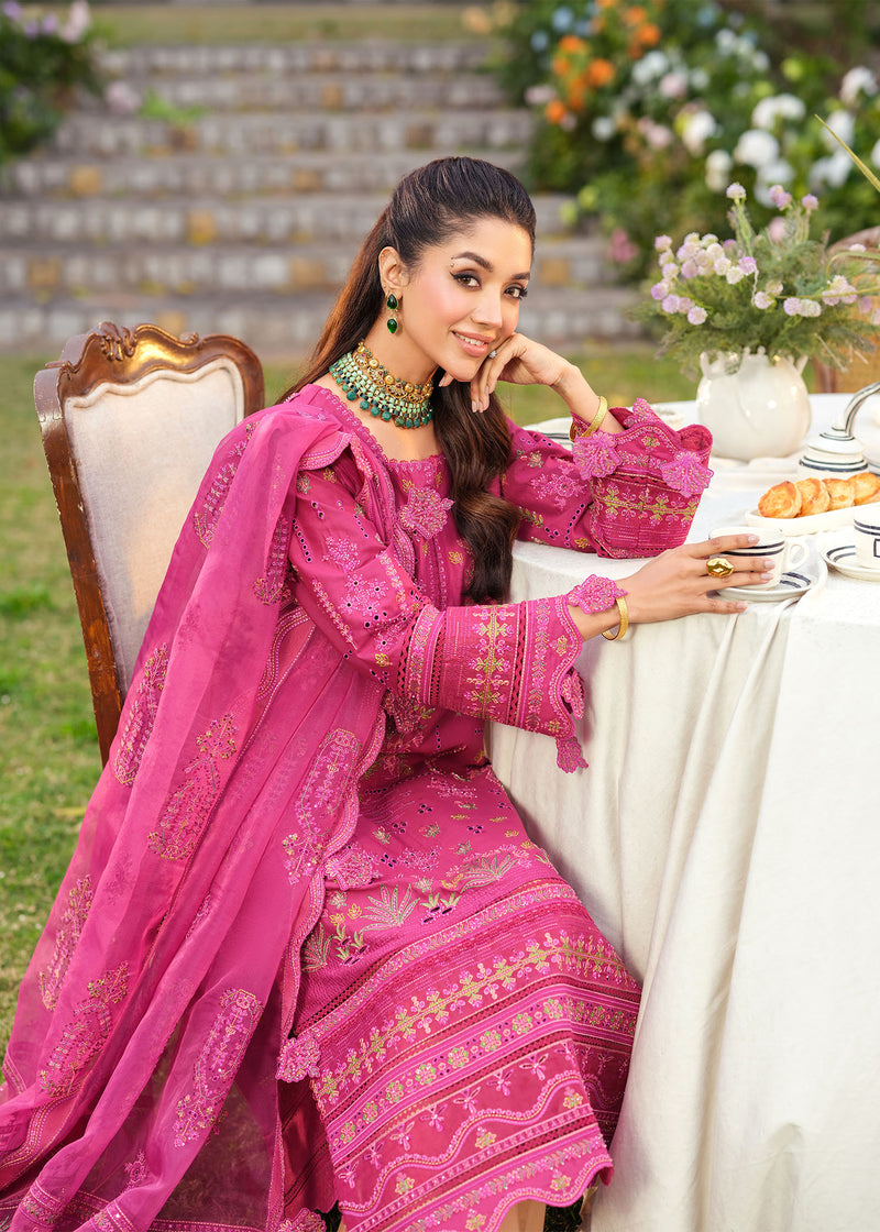 Woman in a pink traditional outfit sitting at a table outdoors with flowers and a staircase in the background.