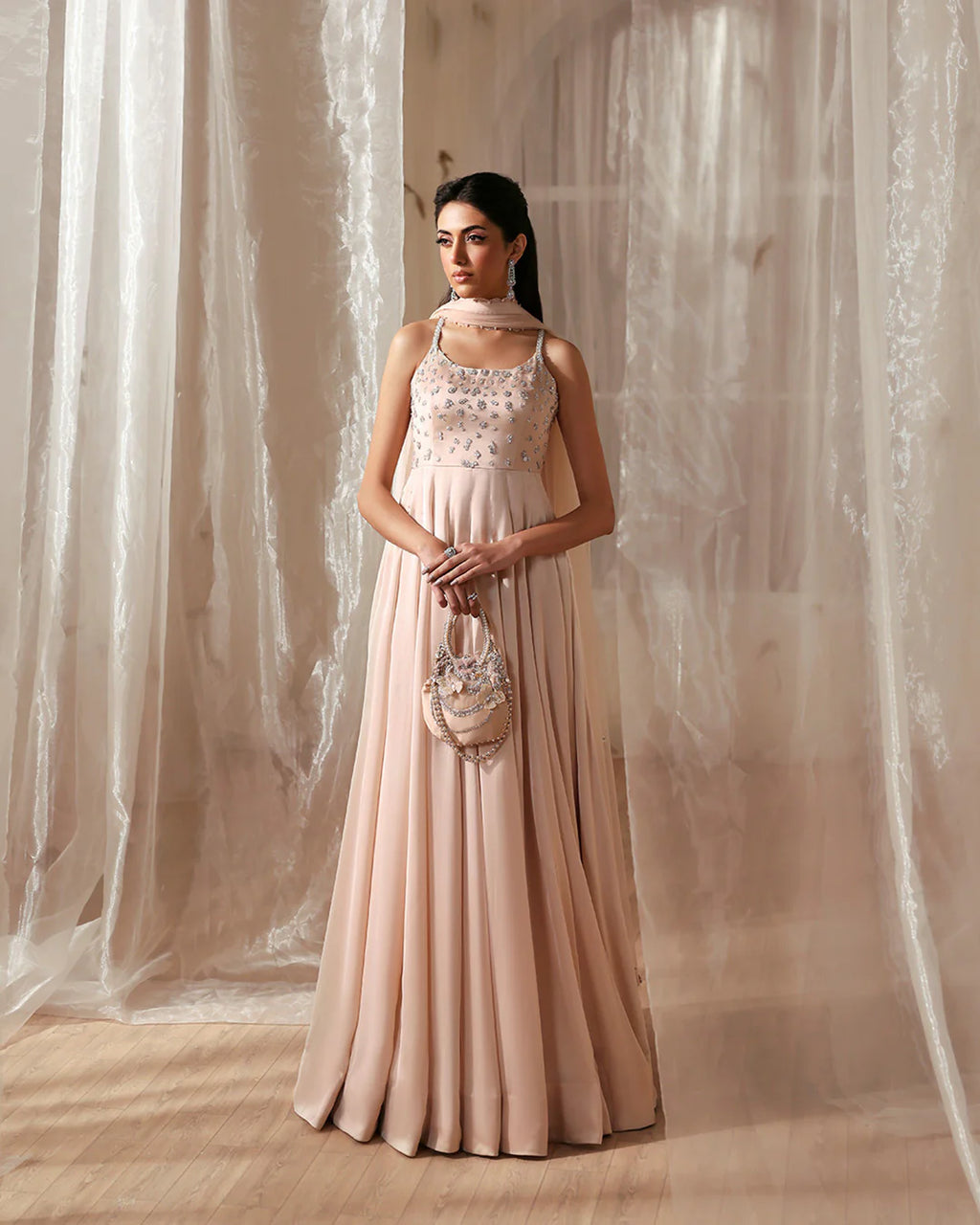 Woman in a long, light pink dress holding a matching handbag against a soft, neutral background.