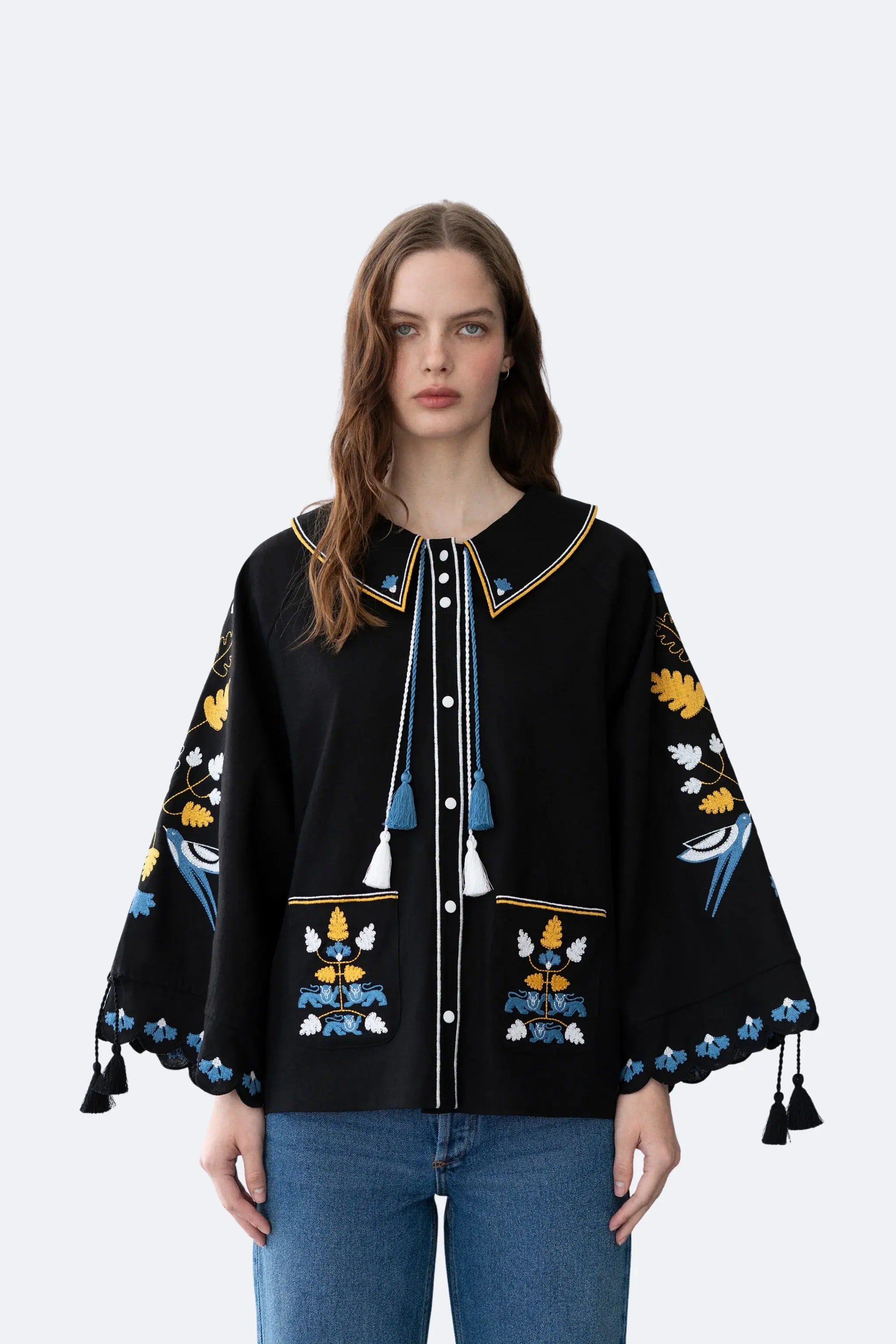 A woman modeling a black embroidered poncho-style top with blue, yellow, and white floral and bird motifs, paired with blue jeans against a light gray studio background.