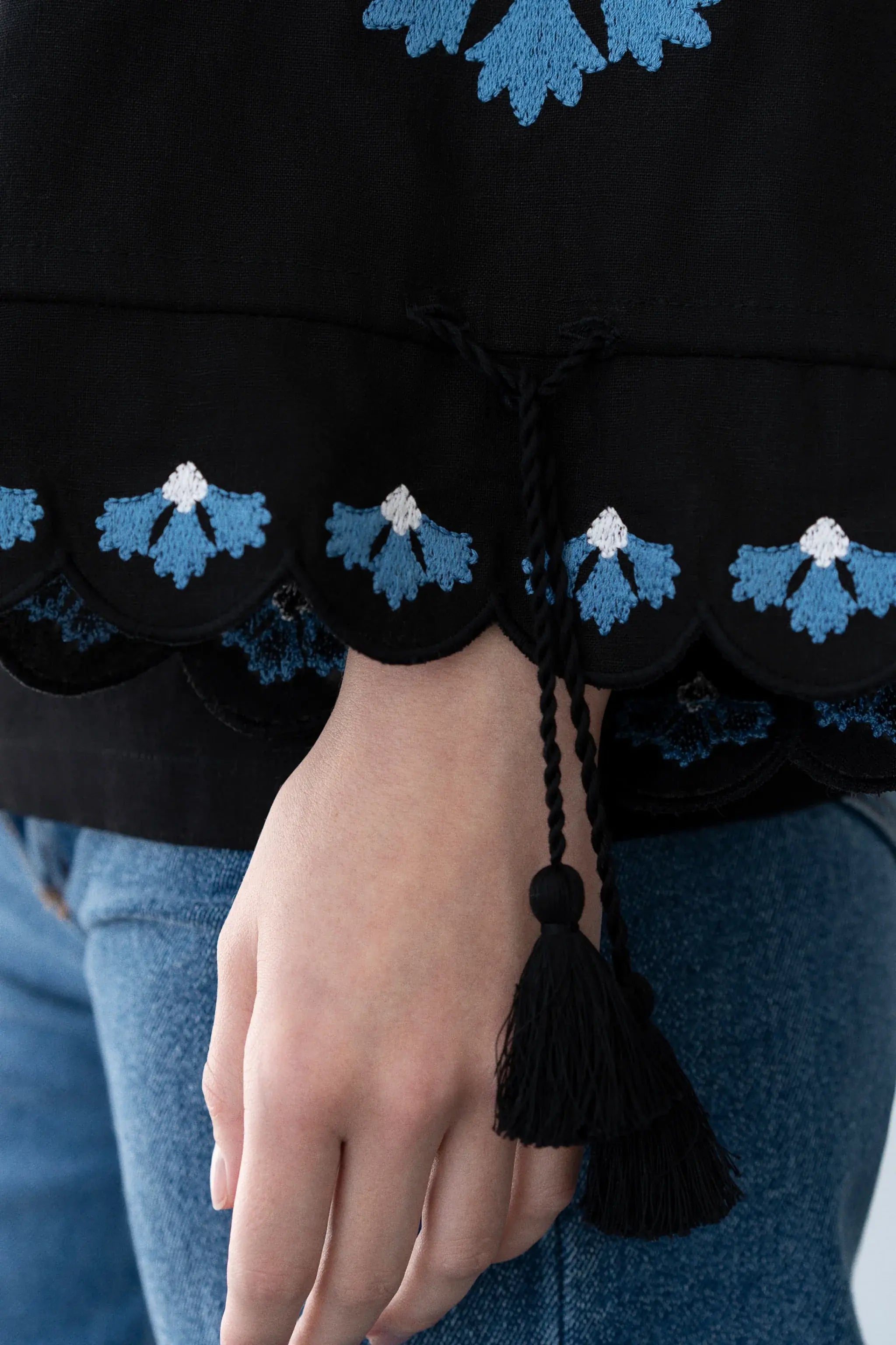 Close-up of a person wearing a black embroidered top with blue and white floral scalloped embroidery, a black drawstring, and a tassel, paired with blue jeans.