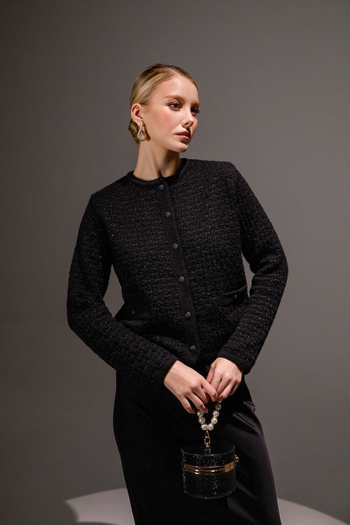 Woman wearing a black textured jacket holding a black handbag against a gray background