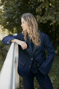 Woman wearing a navy blue blazer standing outdoors with greenery in the background
