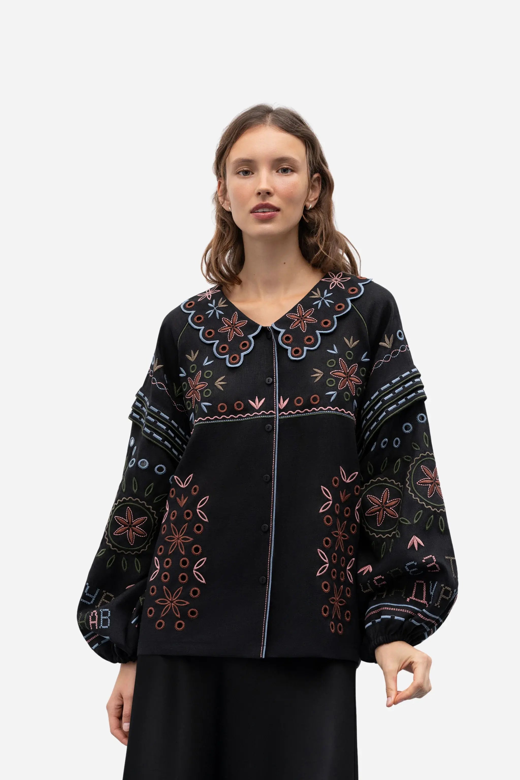 A woman modeling a black embroidered blouse with colorful floral patterns and a scalloped neckline, paired with black trousers, against a plain light gray studio background.