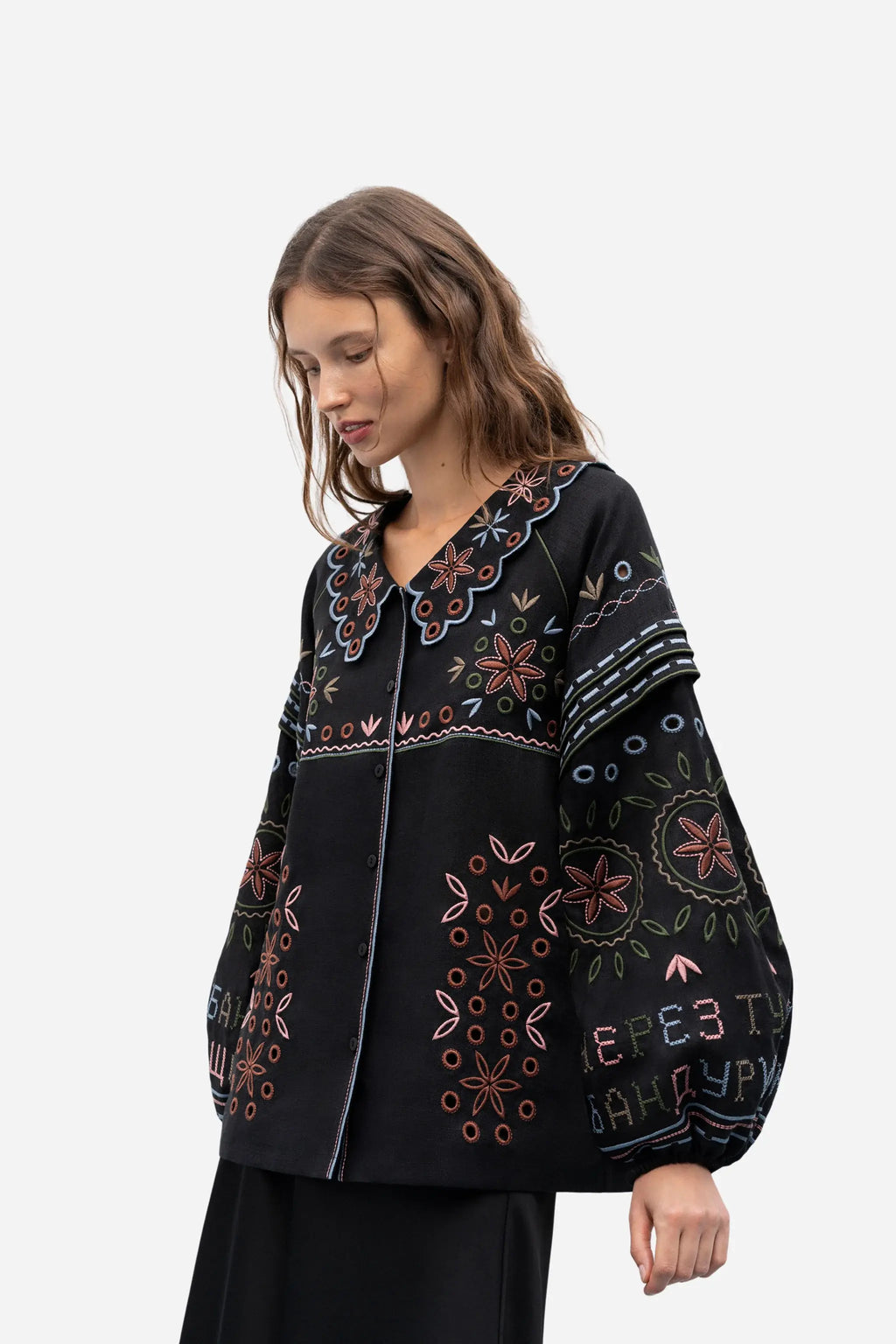 A woman wearing a black embroidered jacket with colorful floral and geometric patterns, photographed against a plain light gray studio background.