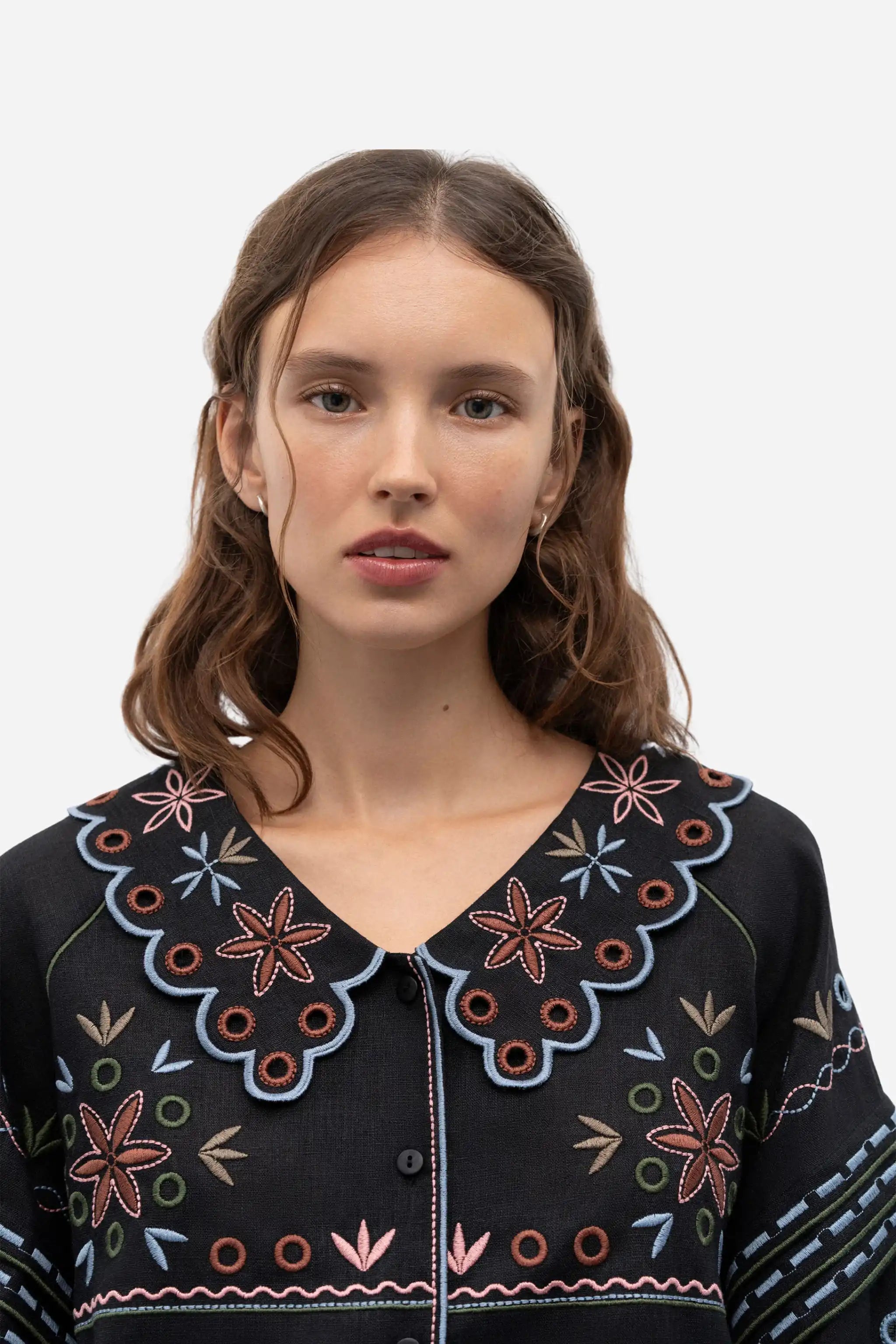 A woman with brown hair wearing a black button-up cardigan with colorful embroidered patterns and a large scalloped Peter Pan collar, photographed against a light gray studio background.