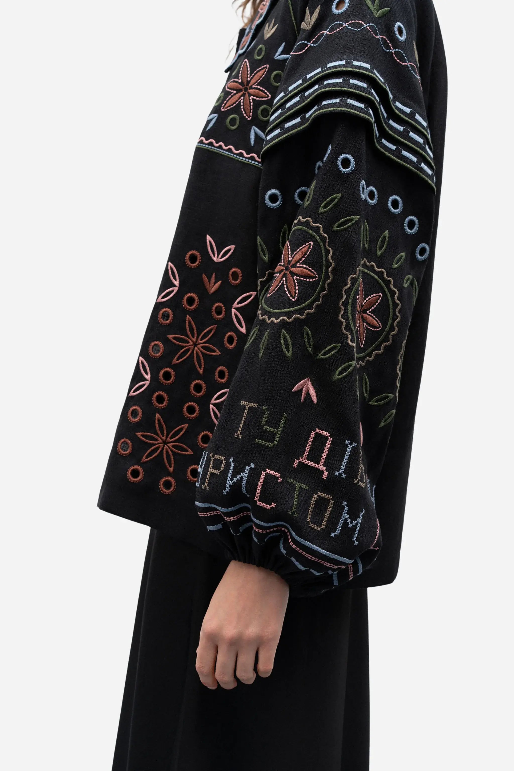 A close-up of a model wearing a black embroidered blouse with colorful floral and geometric patterns and Cyrillic embroidery on the sleeve, paired with a black skirt against a light gray background.