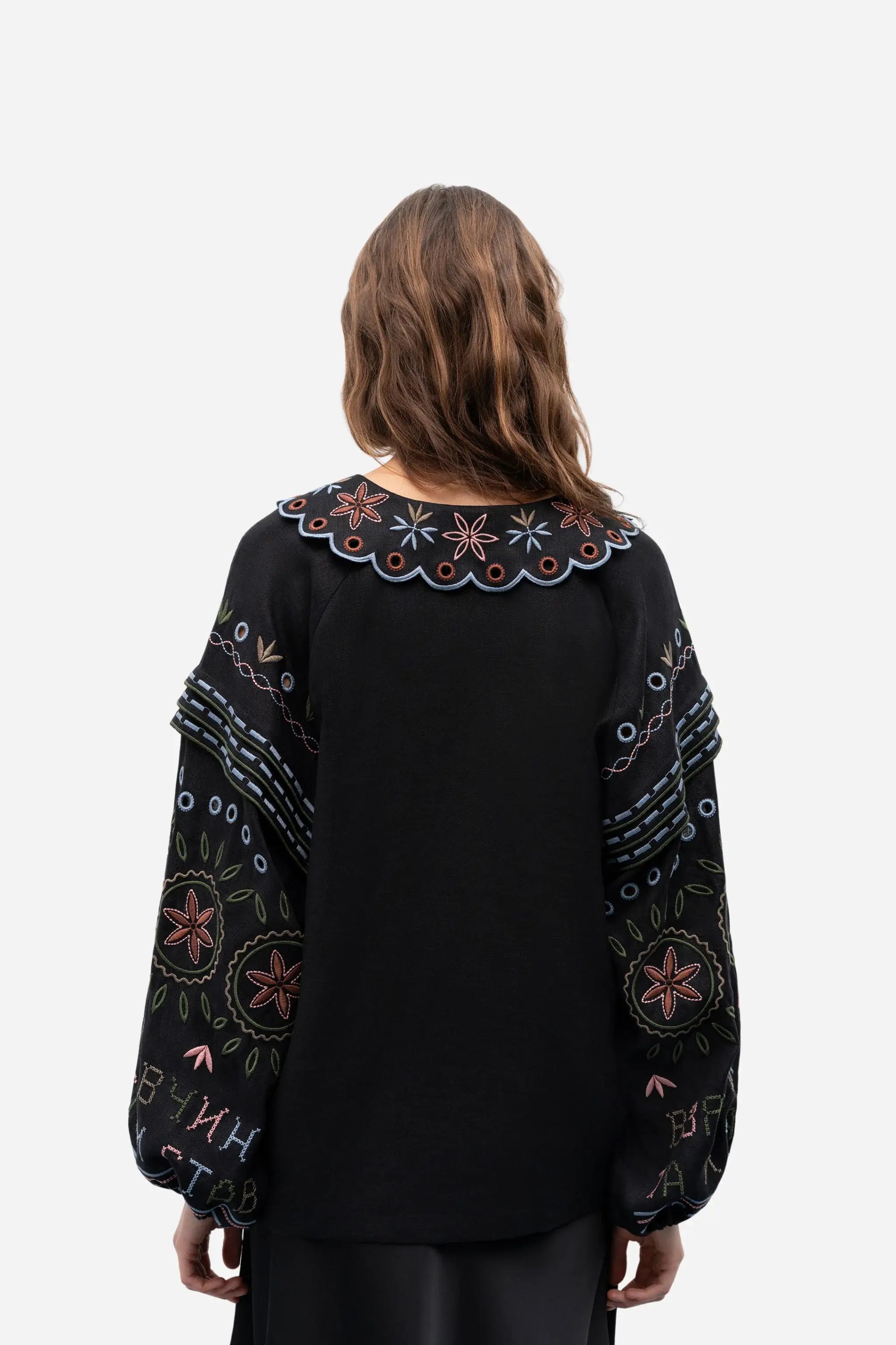 A woman viewed from the back wearing a black long-sleeve blouse with colorful embroidered floral and geometric patterns on the yoke and sleeves, set against a plain light gray studio background.