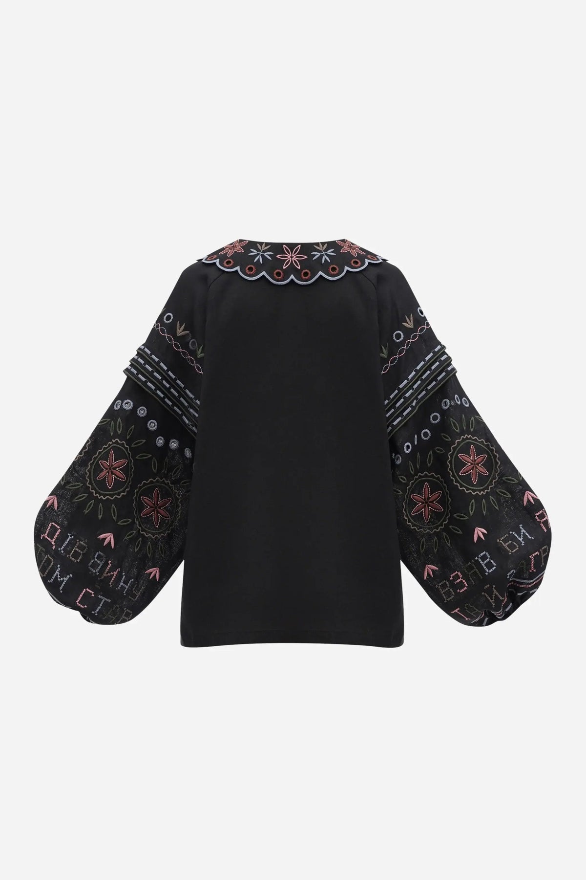 A woman shown from the back wearing a black embroidered blouse with colorful floral and geometric patterns, against a plain light gray studio background.